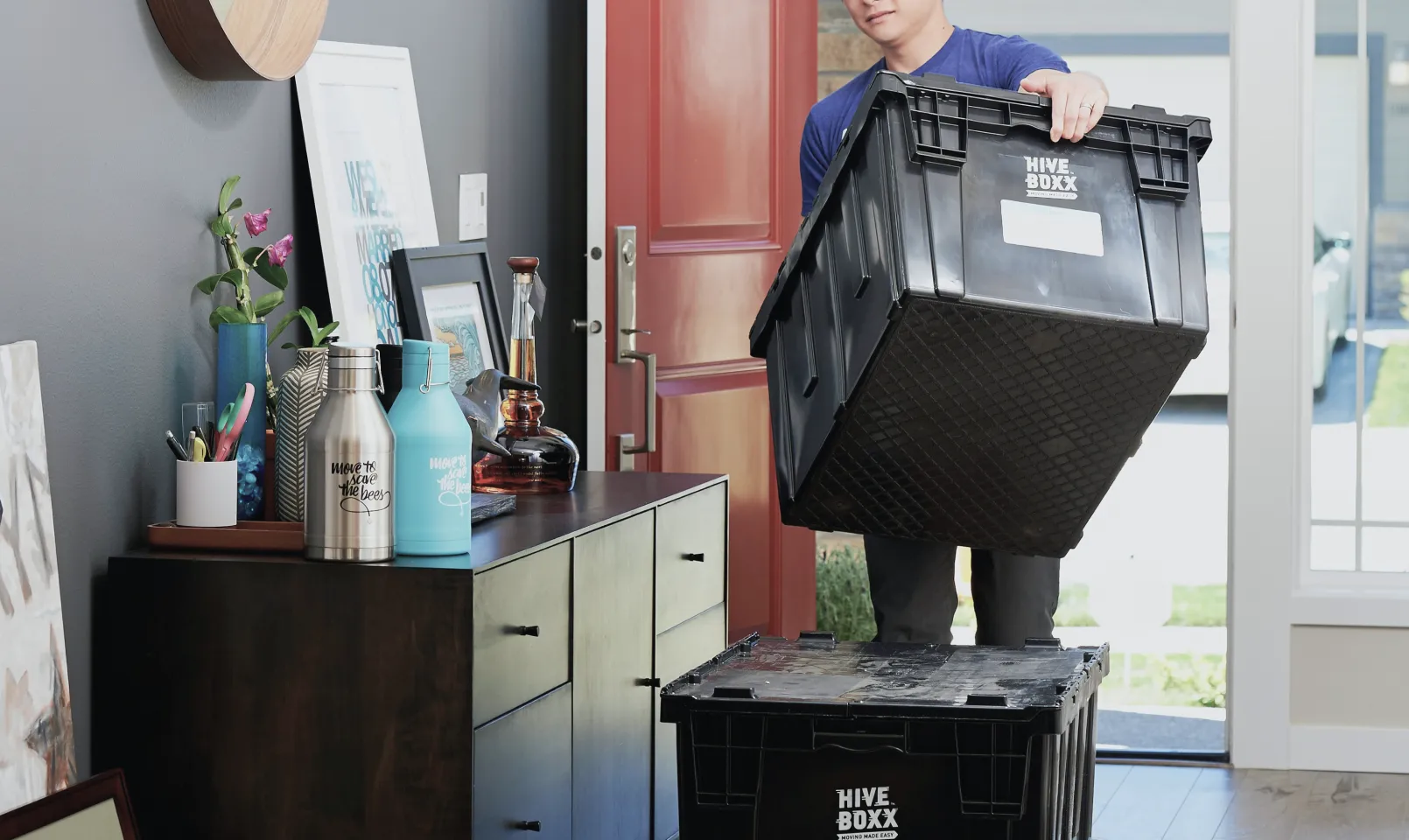 引越し作業中に黒い折りたたみコンテナを運ぶ男性と赤い玄関ドアやキャビネット上の小物